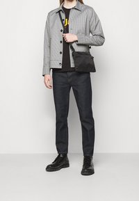 Chaqueta gris a cuadros con botones negros sobre una camiseta negra, combinada con jeans de mezclilla oscuros y botas negras con cordones; llevando un pequeño bolso negro.