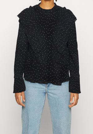 Persoon draagt een zwarte blouse met lange mouwen met witte stippen en ruches, gecombineerd met lichtblauwe spijkerbroek.