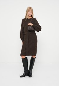 Abito lavorato a maglia marrone con maniche lunghe, vita cintrata, lunghezza al ginocchio. Modella che indossa stivali neri in pelle fino al ginocchio e tiene una piccola borsa marrone.
