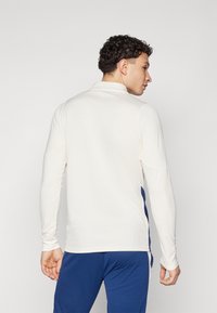 Polo a maniche lunghe di colore crema, realizzato in un tessuto testurizzato, con dettagli in blu navy lungo i lati e colletto pieghevole.