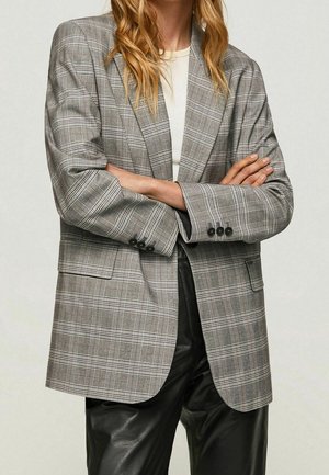 Femme aux bras croisés portant un blazer à carreaux gris, un haut blanc et un pantalon en cuir noir sur un fond uni.