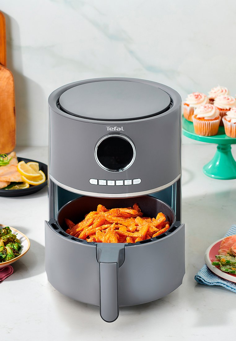 Friteuse à air Tefal grise avec des frites de patates douces dans le panier sur un plan de travail de cuisine entouré de divers plats.