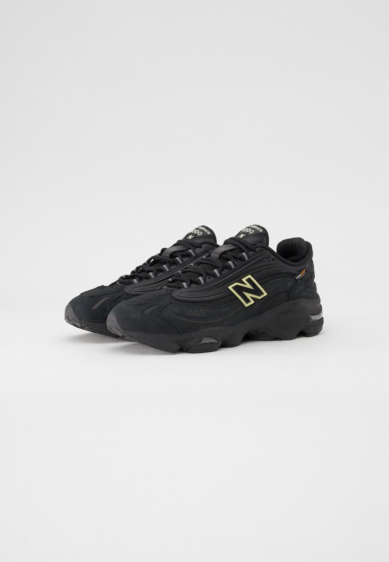 Scarpe da ginnastica nere con una parte superiore testurizzata, caratterizzate da un audace logo "N" giallo, suola in gomma nera spessa e chiusura con lacci.