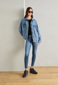 Giacca in denim azzurro chiaro, vestibilità rilassata, con tasche; top aderente nero, jeans skinny blu; sneakers nere con texture liscia e lacci.