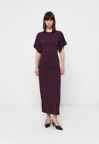 Mauve uska maxi haljina s kratkim rukavima u obliku šiške i naglaskom na struku. Nosila uz crne cipele na visoku petu s špicastim vrhom, na običnoj bijeloj pozadini.