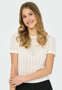 Suéter blanco de punto con mangas cortas, intrincado patrón de encaje y dobladillo acanalado. La modelo lleva un anillo plateado en su mano.