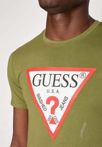 Alyvuogių žalia medvilninė marškinėliai su dideliu raudonu ir baltu trikampiu logotipu, kuriame yra užrašas "GUESS U.S.A." ir klausimo ženklas centre.