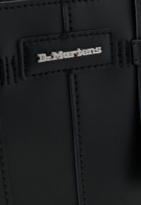 Μαύρη δερμάτινη τσάντα με λαμπερό ασημένιο λογότυπο σε ορθογώνιο πατς, με ορατή ραφή και λεία υφή.