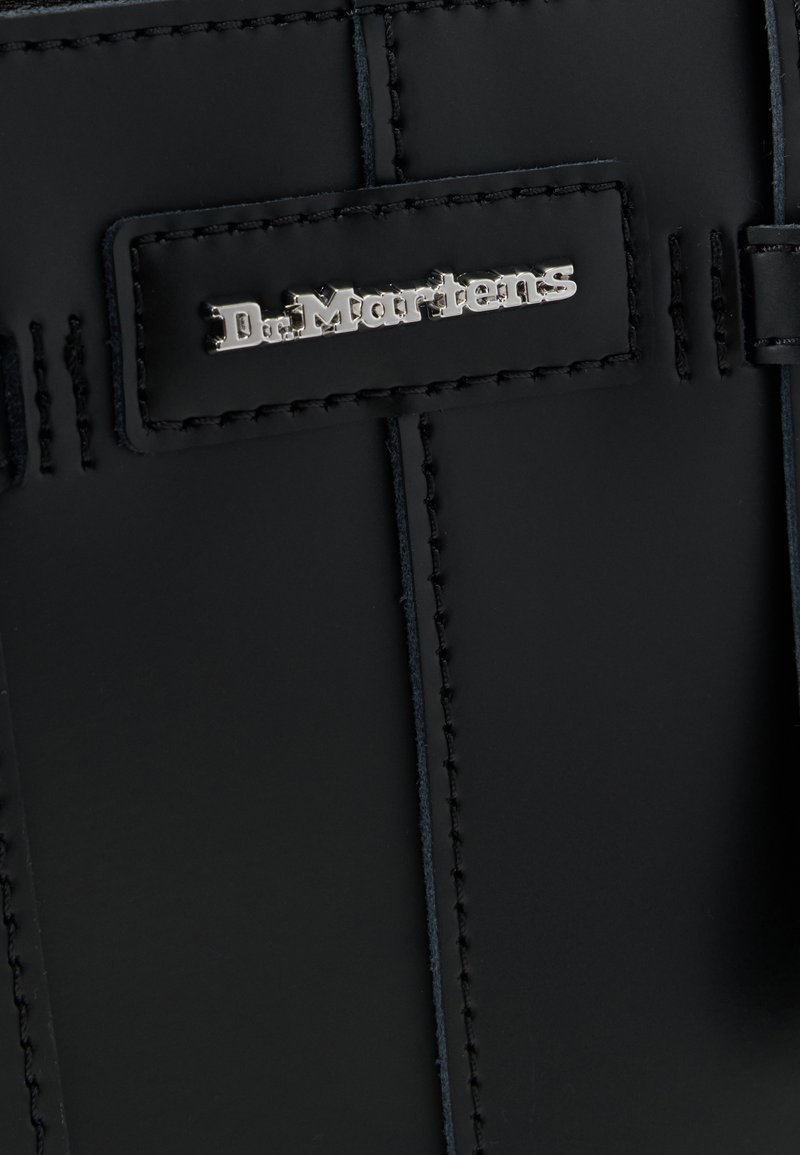 Sac en cuir noir avec un logo argenté brillant sur une étiquette rectangulaire, coutures visibles et texture lisse.