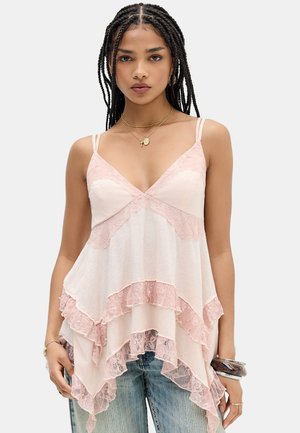 Giovane donna con capelli intrecciati che indossa un top rosa senza maniche con balze e bordatura in pizzo e jeans azzurri, accessoriata con collane e bracciali.