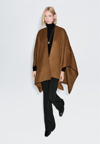 Brun ullponcho med en draperad design, bärs över en svart polokrage. Modellen matchar den med svarta byxor och eleganta, spetsiga skor.