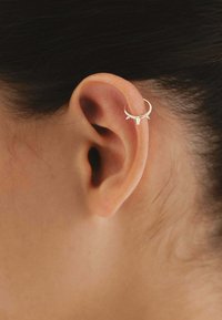 Pendiente de aro dorado con tres picos puntiagudos, llevado en la parte superior de la oreja. Acabado metálico suave y forma circular.