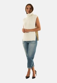 Maglione senza maniche a collo alto a coste bianche con spacco laterale, abbinato a jeans azzurri chiari con risvolto e tacchi alti neri.