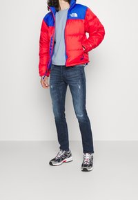 Doudoune rouge et bleue avec un design matelassé, t-shirt bleu clair, jeans usés bleu foncé et baskets grises avec des accents rouges.