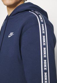 Tumši zila kapuce, kas izgatavota no kokvilnas maisījuma. Tai ir neliels balts Nike logo uz krūšu daļas un vertikāls balts Nike logo svītra uz piedurknes.