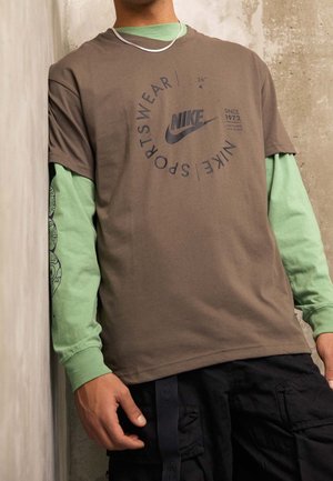 Homme portant un t-shirt de sport Nike marron par-dessus un sous-pull vert à manches longues, pantalon cargo noir, debout devant un mur texturé.