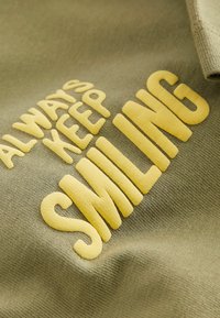 Olivenfarbener Stoff mit erhabenem gelbem Text, der "BLEIB IMMER LÄCHELND" lautet. Eine Nahaufnahme hebt die weiche Textur und die Schriftzügen hervor.