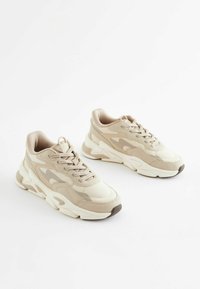 Zapatillas beige con materiales de malla y sintéticos, diseño de cordones, punta redondeada y suela gruesa, con sutiles acentos tonales.