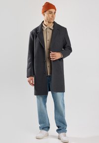 Cappotto di lana grigio con rever a punta, indossato sopra una camicia beige con bottoni, abbinato a jeans blu chiaro e a un berretto di lana color ruggine.