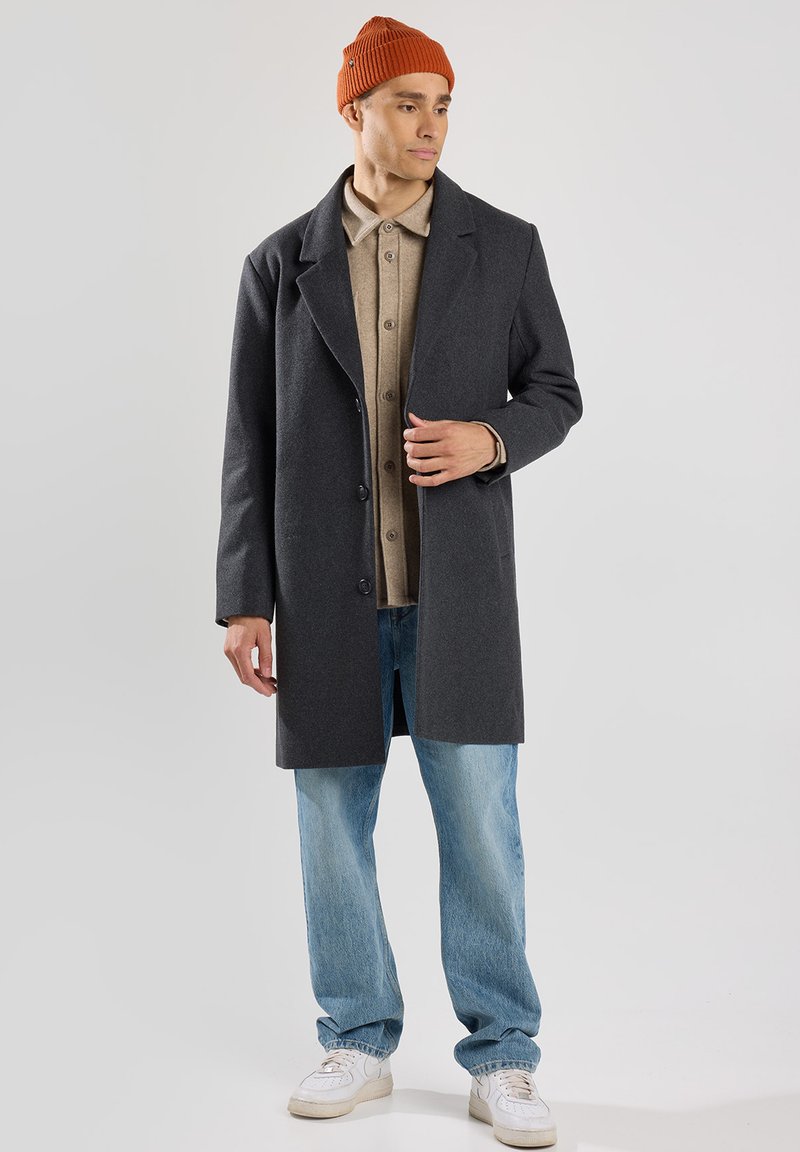 Cappotto di lana grigio con rever a punta, indossato sopra una camicia beige con bottoni, abbinato a jeans blu chiaro e a un berretto di lana color ruggine.