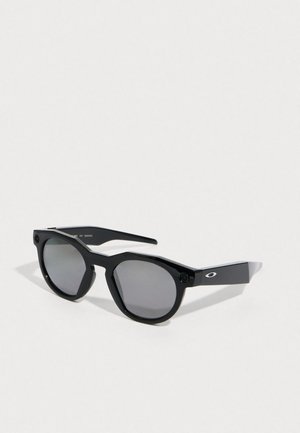 Black sunglasses with a geometric frame, tinted gray lenses, and a textured matte finish. Features distinct side accents and hardware details.