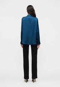 Γυναίκα που στέκεται με την πλάτη προς τα εμάς, φοράει μπλε σατέν μπλούζα με μακριά μανίκια, μαύρο ίσιο παντελόνι και μαύρα ψηλοτάκουνα παπούτσια σε λευκό φόντο.