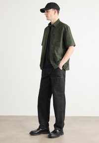 Groene kortekool mouw shirt, zwart t-shirt eronder, donkere spijkerbroek en zwarte instappers. Accessoires omvatten een zwarte pet. Minimalistisch ontwerp.