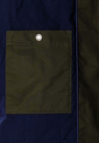 Tela acolchada azul marino con un gran bolsillo de solapa verde oliva, asegurado con un botón de presión plateado, detallado con costuras visibles.