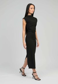 Robe noire sans manches avec un col haut, fente sur le côté et design ajusté. Le mannequin porte des talons noirs à lanières et des bijoux en argent.