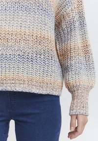 Stickad tröja med en blandning av kräm, orange och blå ränder, med ett texturerat mönster och ribbade muddar. Bärs med mörkblå jeans.