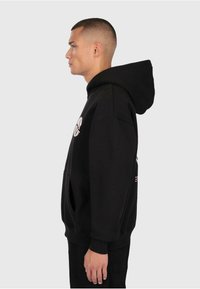 Giovane uomo con capelli corti che indossa un hoodie nero con tasca frontale, mostrato di profilo su uno sfondo chiaro e semplice.