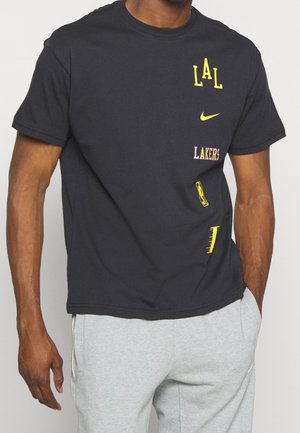 Mężczyzna ubrany w czarną koszulkę z żółto-fioletowymi logo Los Angeles Lakers i Nike, połączoną z jasnoszarymi spodniami dresowymi.