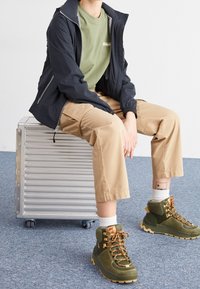 Green hiking boots with orange laces, beige cargo pants, a light green t-shirt, and a black jacket, sitting on a silver rolling case.