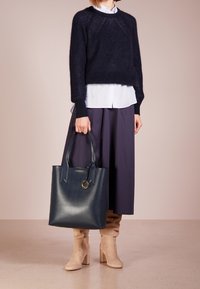 Marineblauer Strickpullover mit rundem Ausschnitt, weißes Hemd mit Kragen, marineblauer plissierter Midirock, beige kniehohe Stiefel mit Absatz und große blaue Tragetasche.