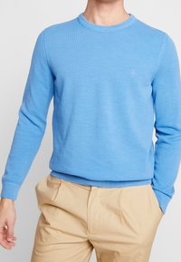 Pull tricoté bleu clair avec un col rond et des poignets côtelés. Texture discrète, avec un petit logo sur la poitrine. Associé à un pantalon beige.