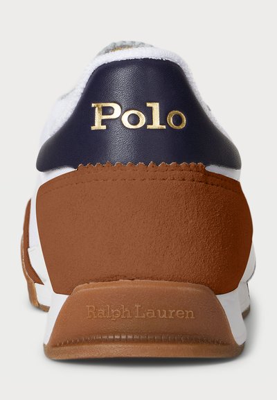 Baltas sneakers ar tumši zilu ādu un brūnu zamšu akcentiem. Uz papēža ir zelta krāsā izšūts vārds "Polo", bet uz zoles ir iespiests "Ralph Lauren".