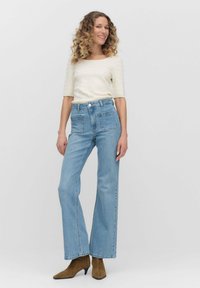 Lichtblauwe high-waisted jeans in een relaxte pasvorm, met voorzakken, gecombineerd met een crème textuur top en bruine puntige enkelboots.