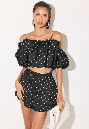Femme en crop top noir épaules dénudées et shorts à pois assortis tenant une pochette blanche, portant de grosses boucles d'oreilles créoles et un maquillage naturel.
