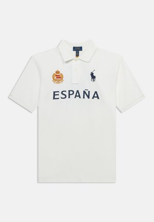 Biele polotričko s golierom, s textom "ESPAÑA", zlatým erbom na ľavej strane hrudníka a tmavomodrým logom hráča póla na pravej strane hrudníka.