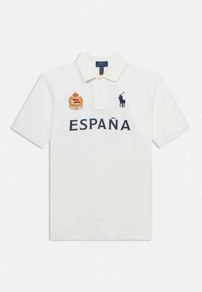 Λευκό πόλο με γιακά, με κείμενο "ESPAÑA", χρυσό έμβλημα στο αριστερό στήθος και λογότυπο παίκτη πόλο σε ναυτικό μπλε στο δεξί στήθος.