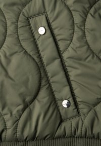 Dark green quilted fabric with two silver snap buttons on a vertical rectangular flap, and ribbed edge at the bottom.