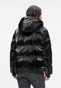 Person von hinten zu sehen, der eine glänzende schwarze Pufferjacke mit Kapuze und dunkelgraue Jeans trägt, vor einem schlichten hellen Hintergrund.