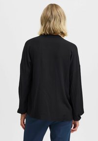 Person med axellångt blont hår som bär en lös svart långärmad topp och mörkblå byxor, vänd bort från kameran mot en heltäckande bakgrund.