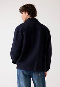 Tengerés kék bolyhos kabát gallérral és oversized szabással, texturált felülettel. Világoskék farmernadrággal párosítva.