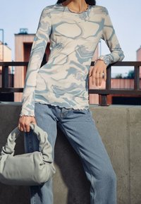 Maglietta a maniche lunghe con un motivo a marmo blu e bianco, tessuto morbido, orlo scallop, abbinata a jeans chiari a vita alta e una borsa grigia.