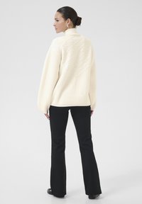 Cream ribbed knit sweater with high collar, featuring a textured diamond pattern on the back; paired with black flared pants.