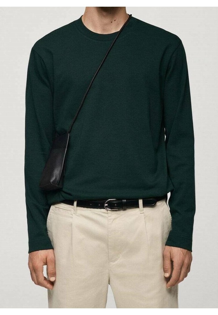 Homme portant une chemise à manches longues vert foncé, un pantalon crème avec ceinture noire, et un petit sac bandoulière noir suspendu à l'épaule.