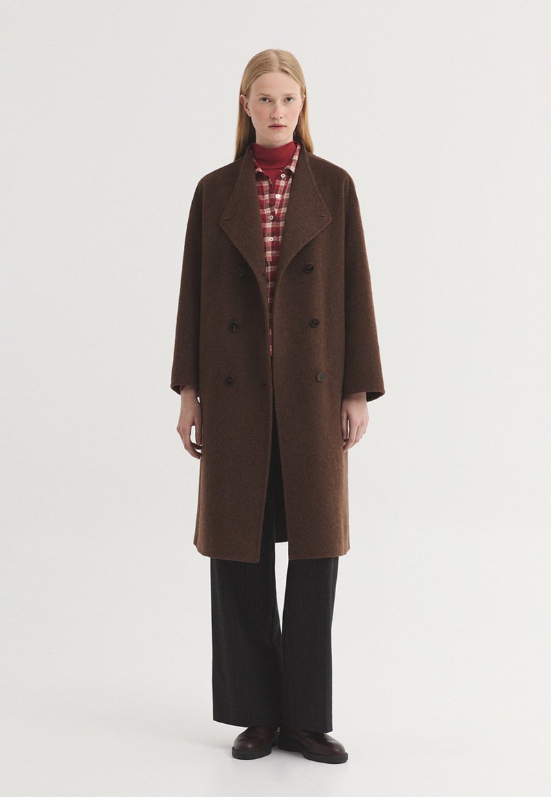 Femme portant un manteau en laine marron à double boutonnage par-dessus une chemise rouge à carreaux et un pantalon noir à jambes larges, debout sur un fond blanc uni.