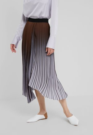 Kobieta ubrana w plisowaną spódnicę o wysokim stanie z gradientem od brązowego do szarego oraz białe wsuwane buty, stojąca na jednolitym tle.