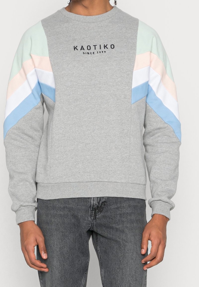Personne portant un sweat-shirt gris avec le texte « KAOTIKO SINCE 1999 » et des rayures chevrons pastel bleu, blanc, rose sur les manches, associé à un jean gris foncé.